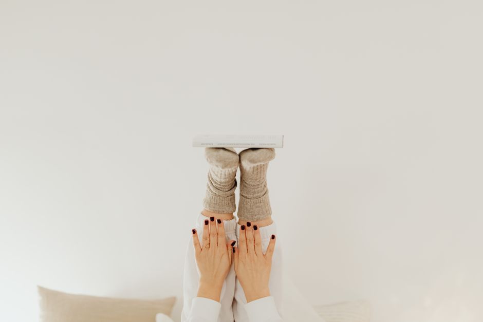 A pair of feet in socks balances a book against a white backdrop, with hands supporting the pose.