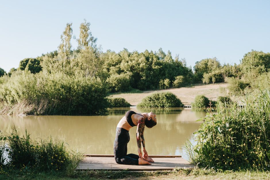 Side view of young barefoot tattooed lady showing Ustrasana pose while practicing yoga on pier near river and bushes