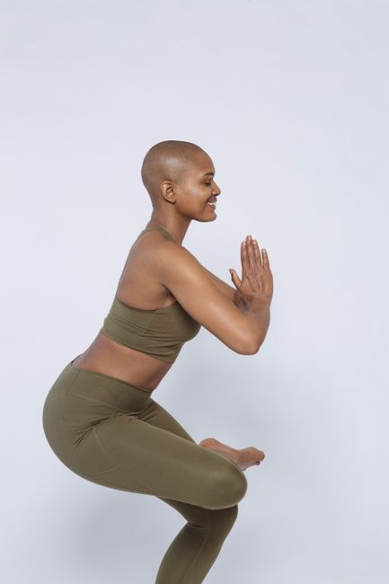 Side view of smiling African American female in khaki sportswear standing in Eka Pada Utkatasana with closed eyes against white background