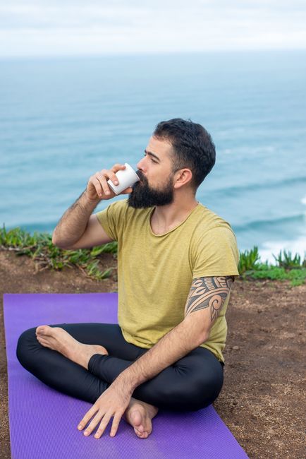 Bearded man with tattoo meditates by the sea in Portugal holding a cup.