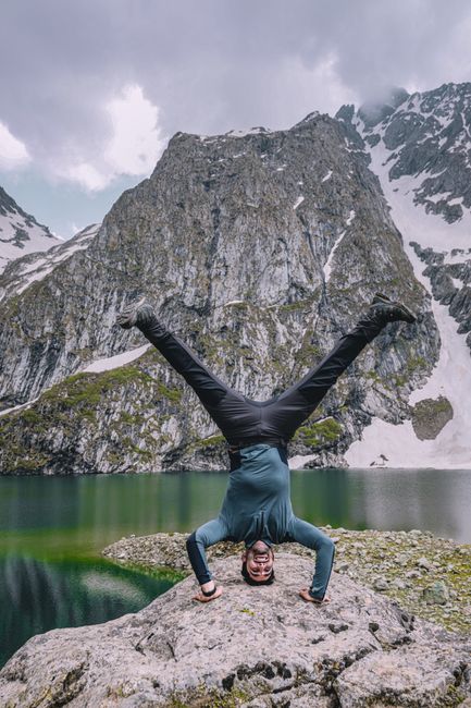 A man performs a headstand on a rock near a lake surrounded by mountains and snow.