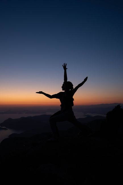Silhouette of a woman with arms raised at sunset on a mountaintop, embodying freedom and tranquility.