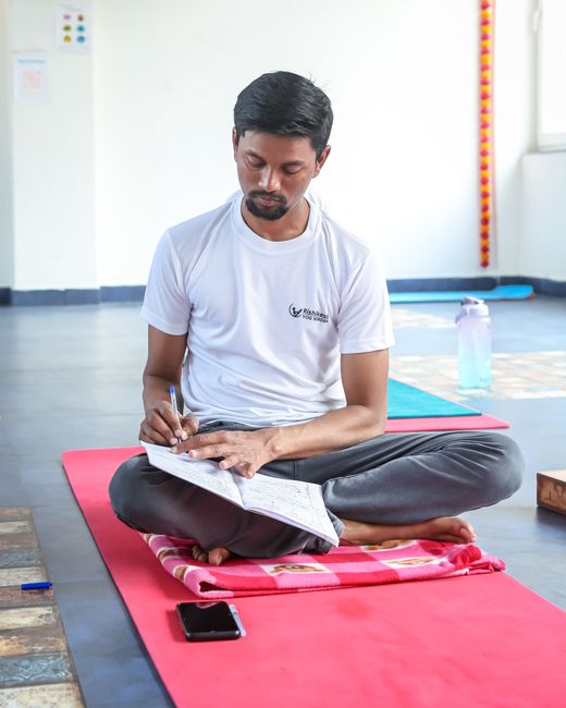 A man sitting cross-legged on a yoga mat, writing in a journal indoors.