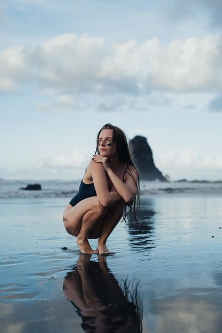 A woman squatting on a beach with stunning reflections and a rock formation in the background.