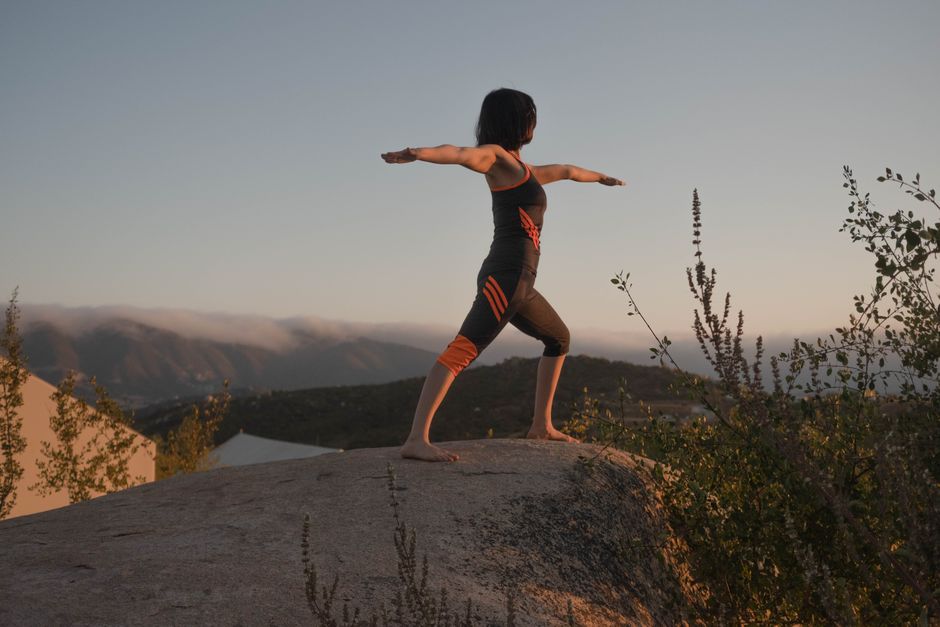 Woman in warrior II yoga pose on a rock at sunset, embracing wellness and mindfulness.