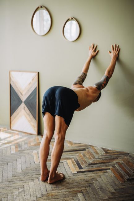 A tattooed man performs a yoga stretch against a wall indoors, showcasing flexibility and balance.