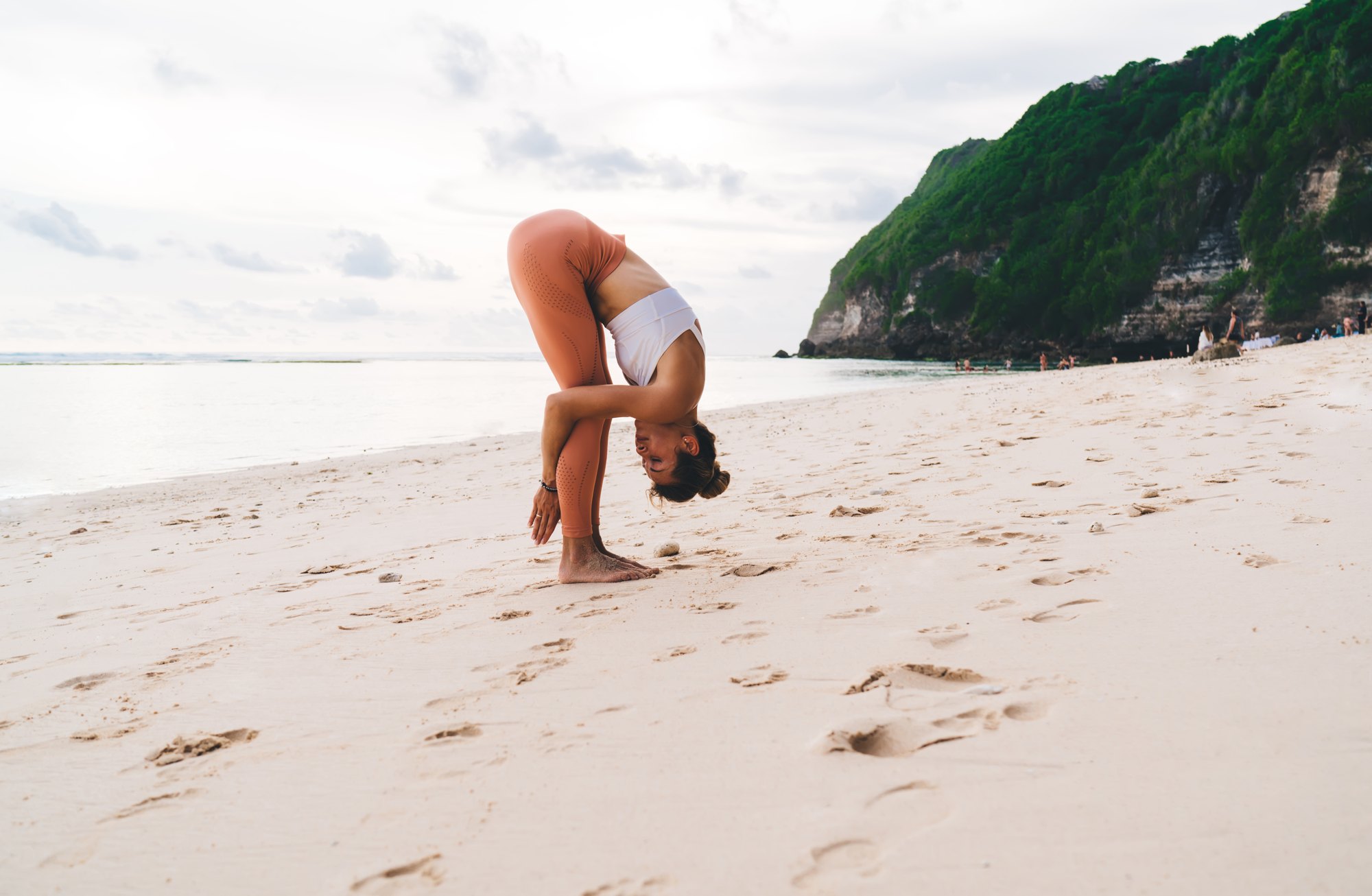 Hands under feet pose on a beach below green cliffs