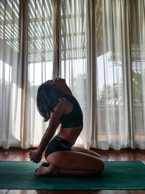 Woman practicing yoga indoors, kneeling on a mat with natural daylight streaming through large windows.