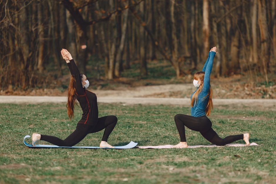 Female friends in sterile masks and sportswear standing in Anjaneyasana pose while practicing yoga on mats in park during COVID 19 pandemic