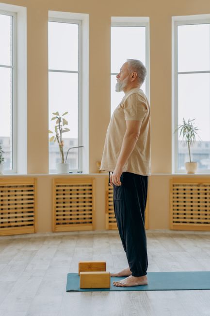 Senior man standing on yoga mat indoors, engaging in mindful exercise with natural sunlight streaming through windows.