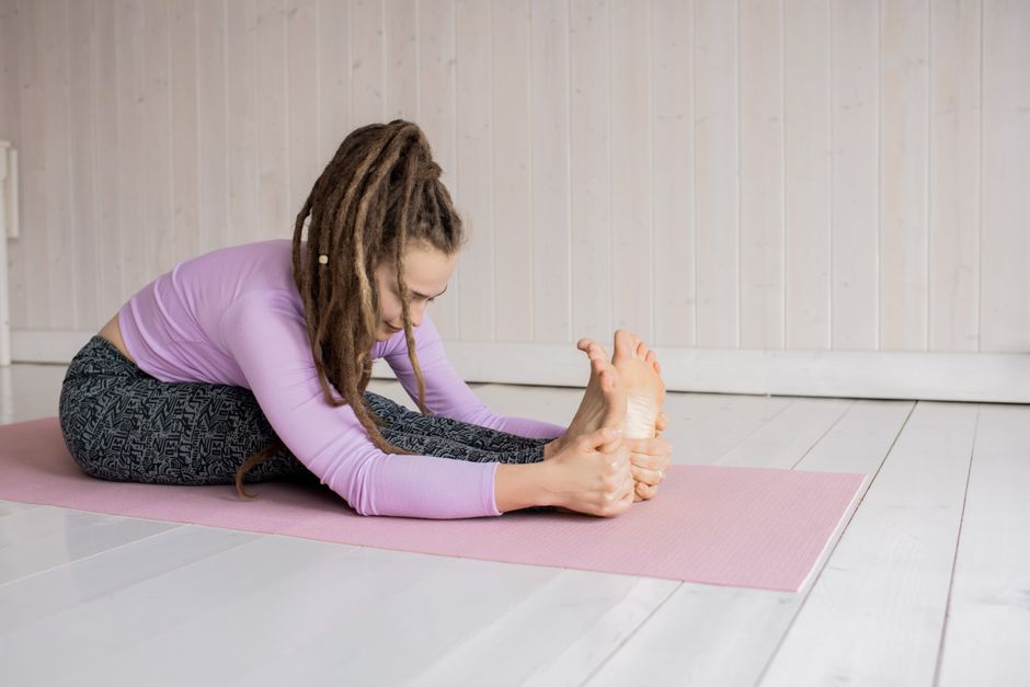 A woman stretching in a seated forward bend yoga pose on a pink mat indoors.