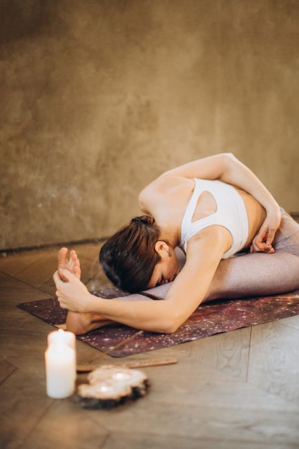 A woman performing seated forward bend yoga pose on a mat indoors. Relaxing ambiance with candles.
