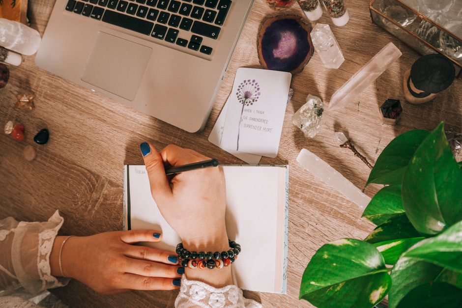 Woman's hand writing in a journal surrounded by crystals, plants, and laptop on a wooden desk.