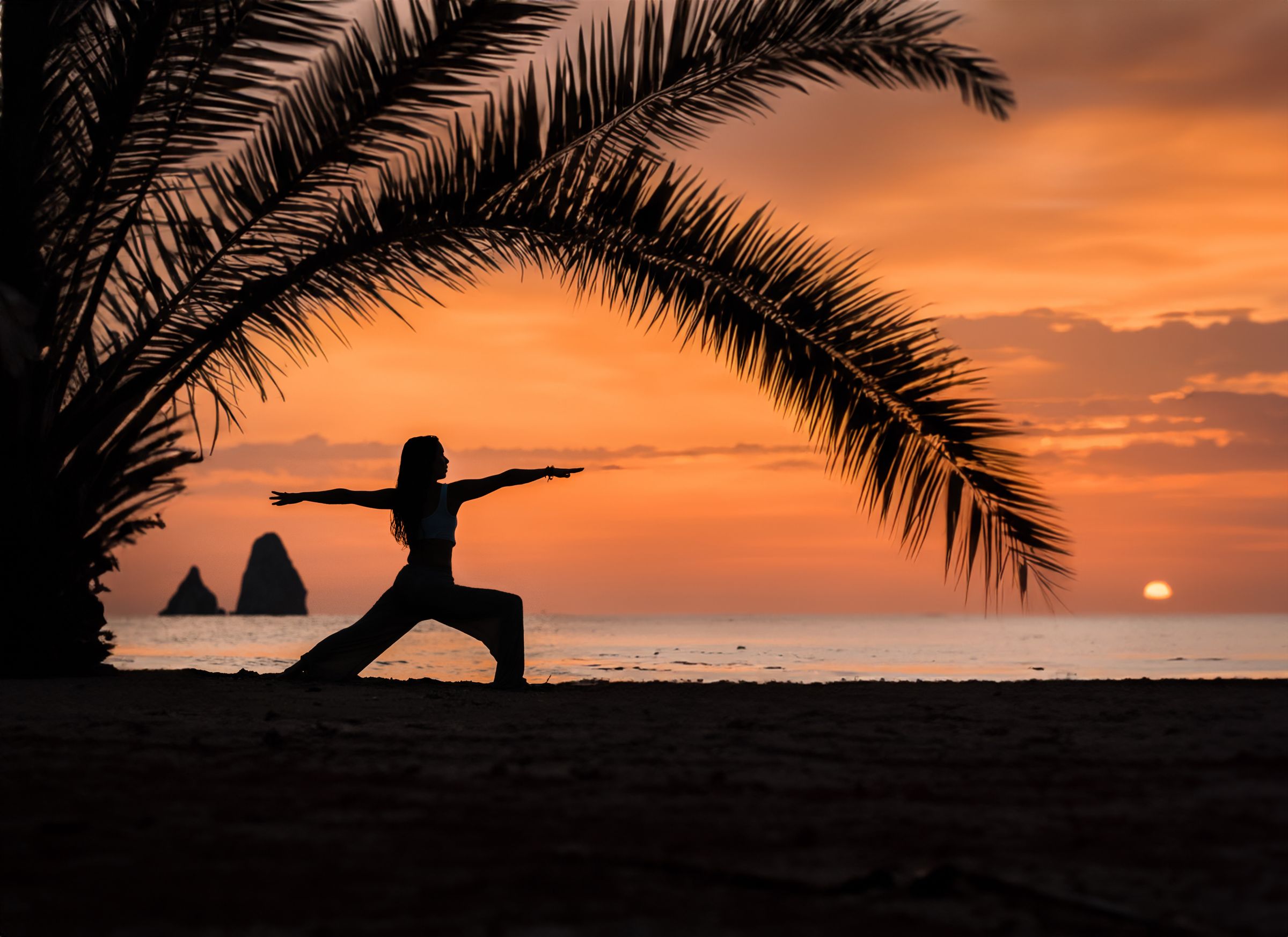 Woman in a yoga pose under palm trees on a beach at sunset