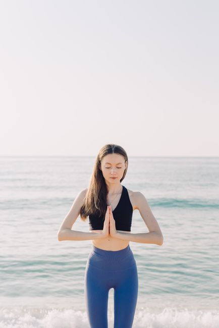 Woman practicing yoga by the ocean, meditating with hands together and eyes closed. Peaceful seascape.