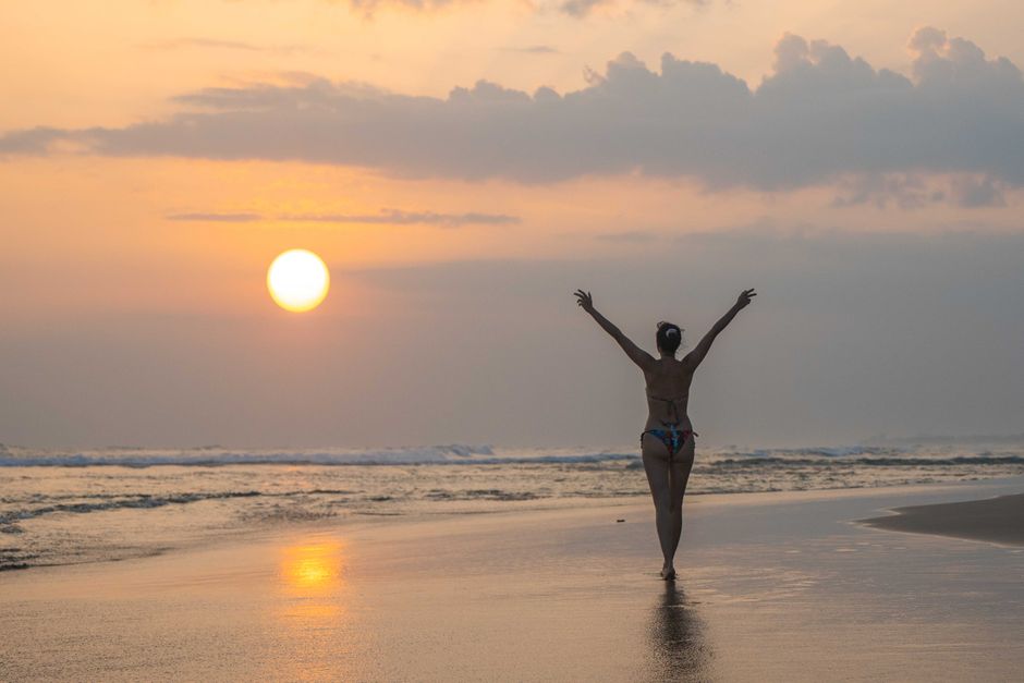 Silhouette of a woman in a bikini facing sunrise on the beach, arms outstretched.