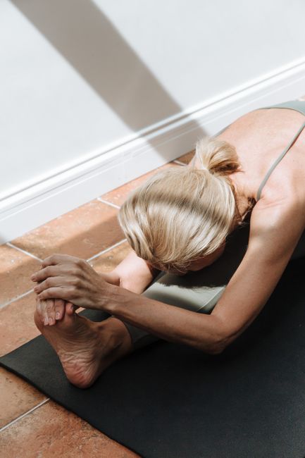 Woman performing a seated forward bend on a yoga mat, indoors in natural sunlight.