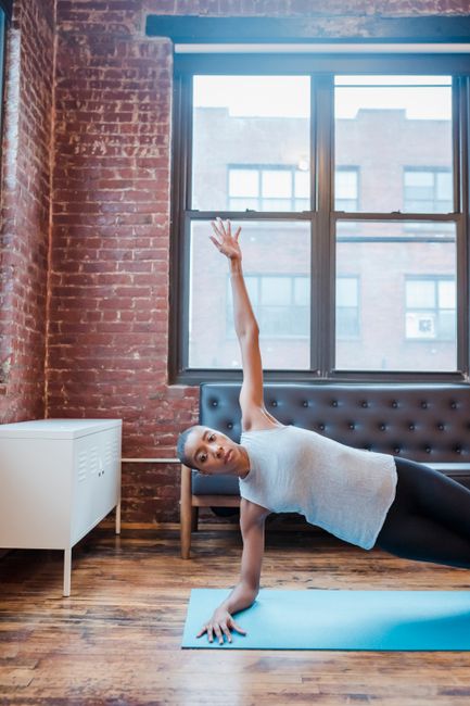 Sportive African American female in active wear doing side plank with raised arm doing yoga training on mat at home