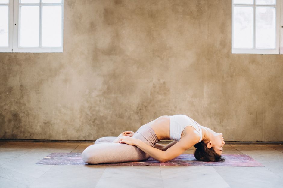 A woman performing Matsyasana yoga pose on a mat indoors, focused on relaxation and wellness.