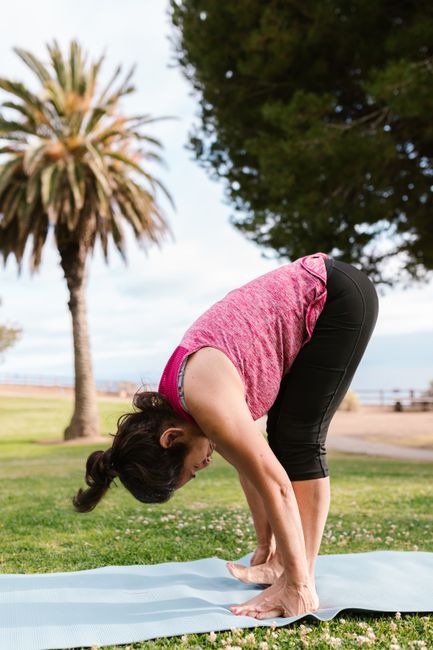Outdoor yoga session with a woman performing a forward bend on a mat, surrounded by nature.