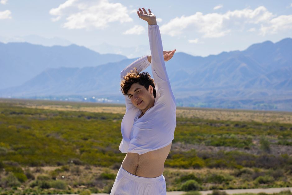 A young man in white clothes stretching outdoors with mountains in the background, showcasing a serene and natural setting.