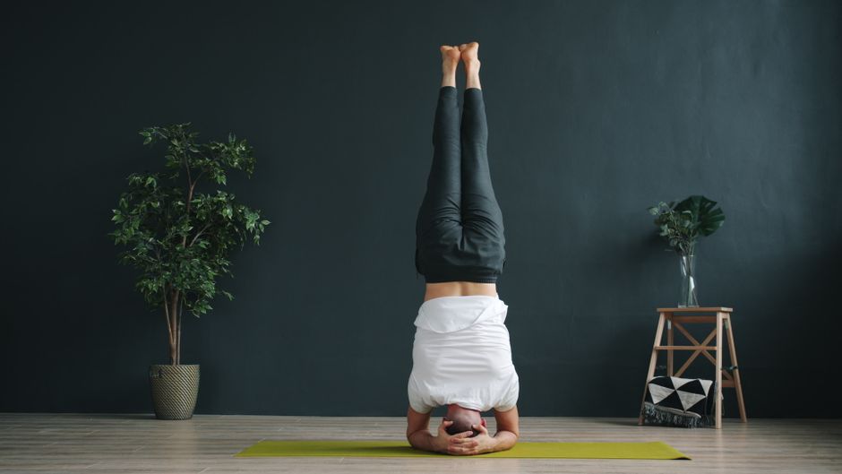 An adult performing a headstand yoga pose on a mat indoors, with plants in the background.