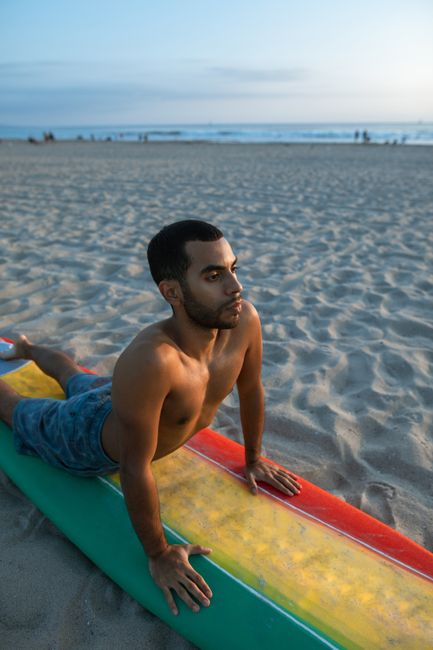A man performs yoga on a colorful surfboard at the beach during sunset.