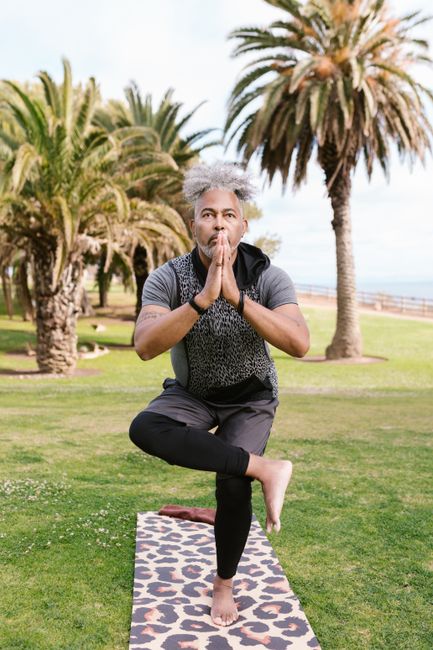 Man in a balancing yoga pose on a mat in a sunny park with palm trees.
