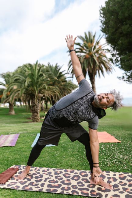 Senior man doing yoga in a park setting, demonstrating triangle pose under palm trees.