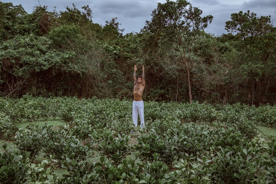 Shirtless man practicing yoga with arms raised in a green field, promoting wellness and mindfulness.
