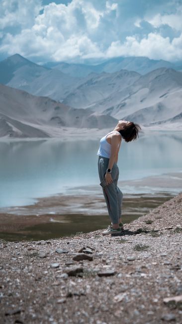 A woman stands bent backward by a tranquil mountain lake in Xinjiang, China, embracing nature's beauty.