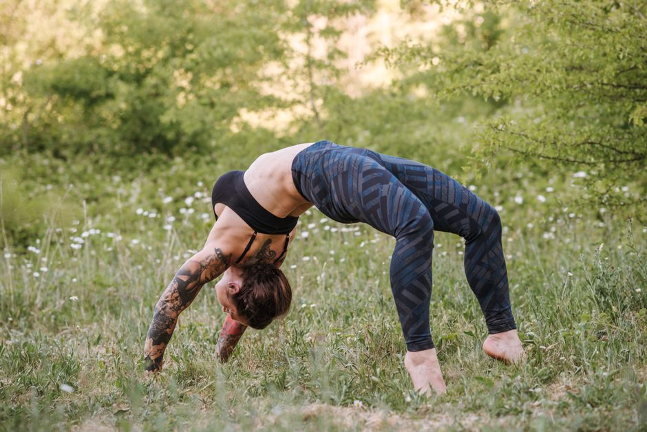Side view of unrecognizable flexible female in sportswear standing in Urdhva Dhanurasana pose while practicing yoga on meadow