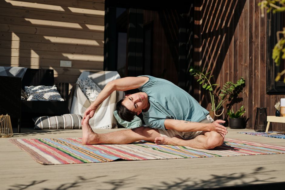 Adult man in yoga pose stretching on a colorful mat on a sunny outdoor veranda.