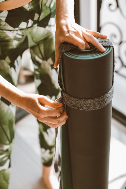 Close-up of a woman rolling up a green yoga mat indoors, with focus on hands and printed leggings.